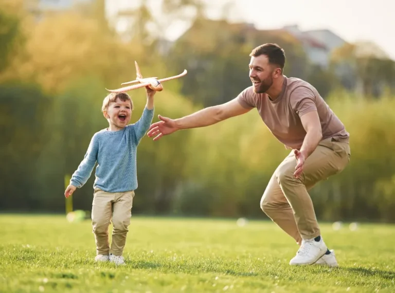 Young boy holding wooden toy airplane running on grass with smiling man reaching out outdoors.