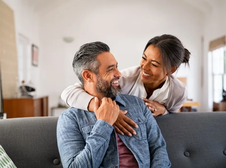 Middle-aged couple smiling and embracing on a gray sofa in a bright living room.